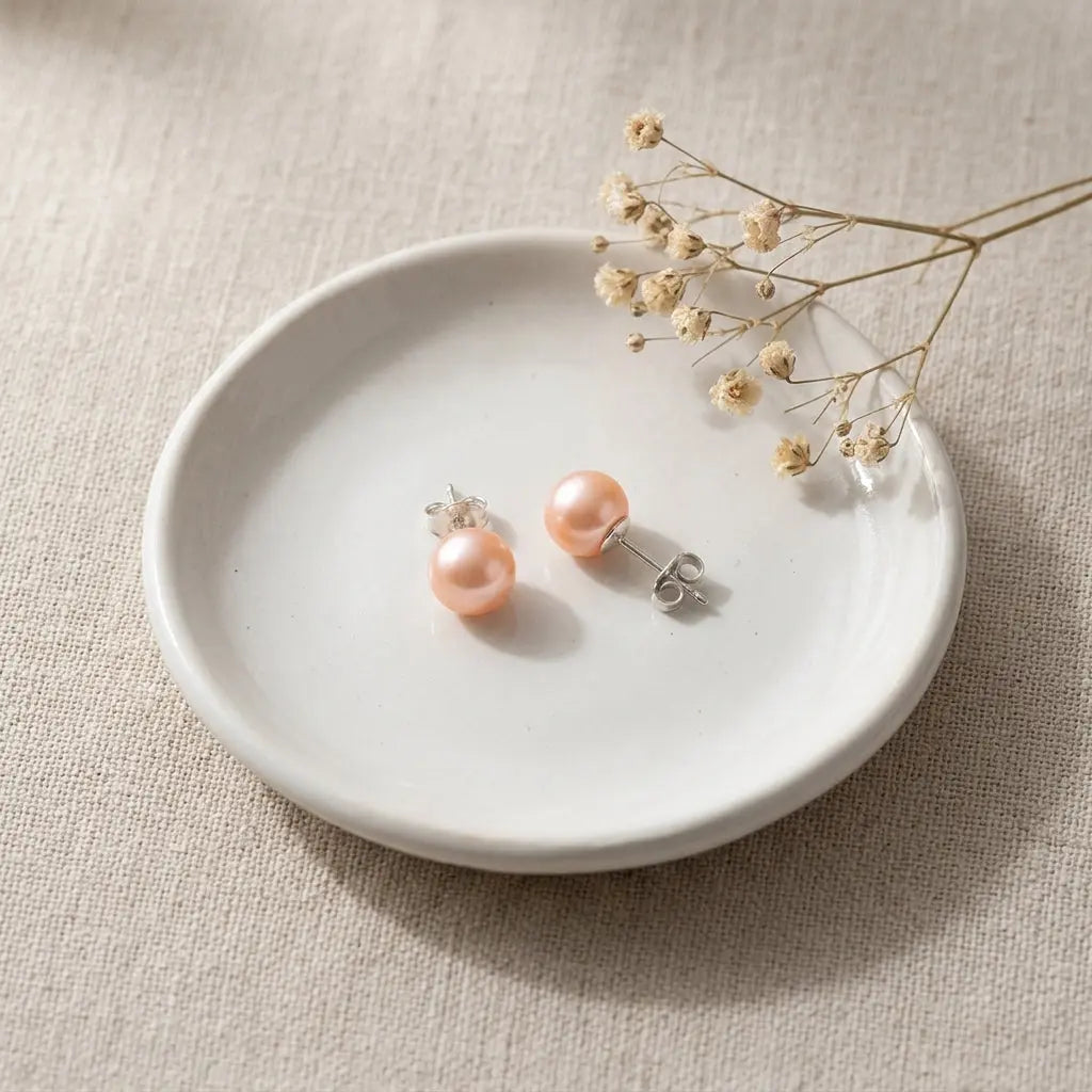 Lustrous peach freshwater pearl studs resting on a minimalist ceramic jewelry dish flat lay
